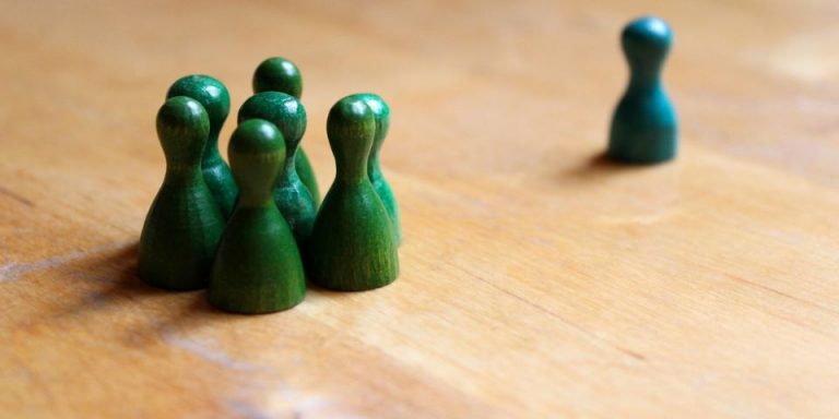 Mikroaggressionen Eine einzelne grün-blaue Spielfigur steht abseits von einer Gruppe grüner Figuren. Das Bild verdeutlicht Ausgrenzung, soziale Isolation und Formen der Diskriminierung.