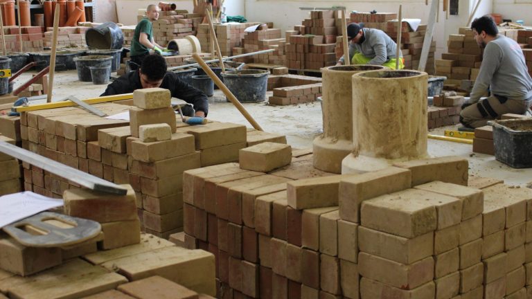 Vier Personen lernen in einer Werkstatt Bauarbeiten. Sie arbeiten mit Ziegeln und verschiedenen Werkzeugen.