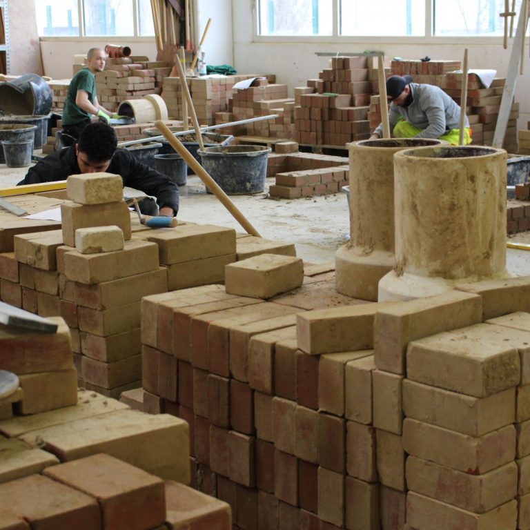Vier Personen lernen in einer Werkstatt Bauarbeiten. Sie arbeiten mit Ziegeln und verschiedenen Werkzeugen.