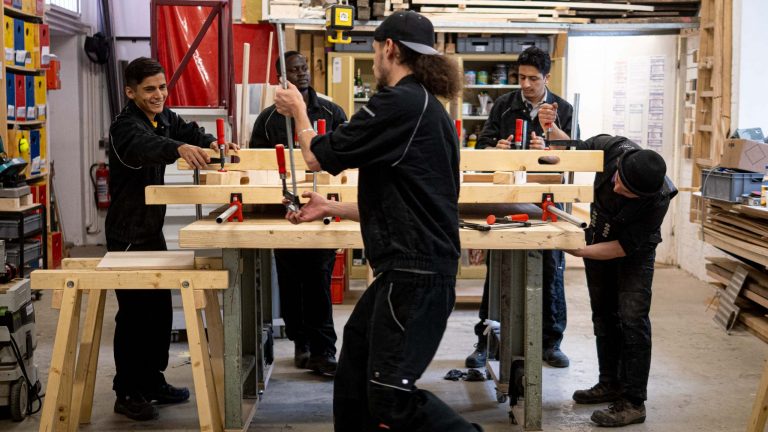 Fünf Personen lernen und arbeiten gemeinsam in einer Holzwerkstatt mit Werkzeugen in einer angenehmen Umgebung.