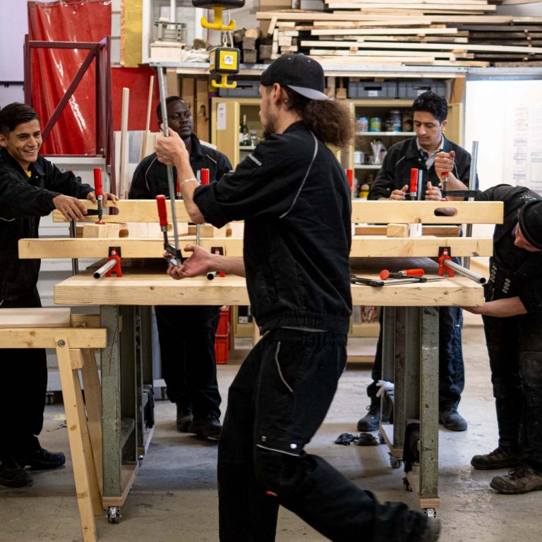 Fünf Personen lernen und arbeiten gemeinsam in einer Holzwerkstatt mit Werkzeugen in einer angenehmen Umgebung.